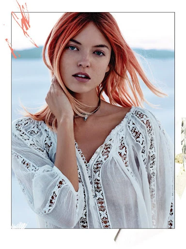 Woman with red hair wears an off-white boho blouse in front of a blurred pale blue sea, posing in a medium shot.
