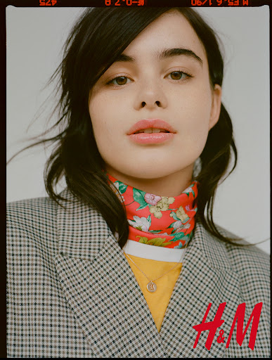 Close-up of a young woman with dark hair wearing a plaid blazer, a yellow shirt, and a red floral scarf against a grey backdrop.