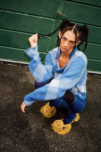 A stylish person with braided hair, wearing a blue sweater, poses outdoors in trendy sandals with a green door in the background.