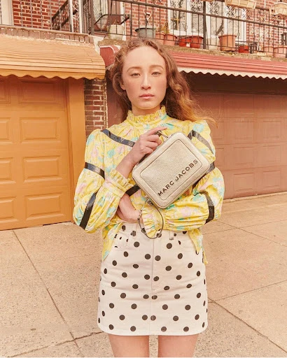Young woman in patterned blouse and polka dot skirt holds a Marc Jacobs purse in an outdoor setting, posing in front of a brick building.