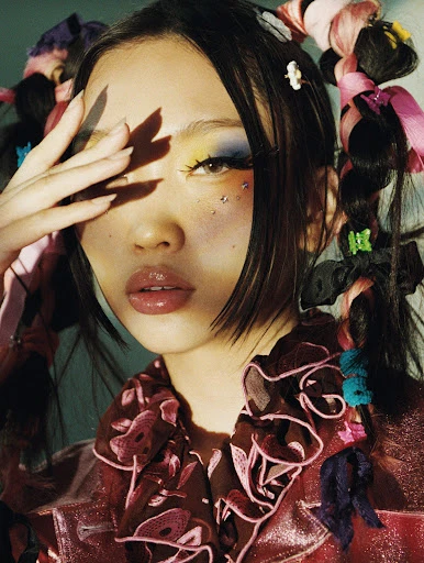 Close-up studio portrait of an Asian woman with colorful eyeshadow, hair accessories, and a patterned shirt, partially covering her face with her hand.
