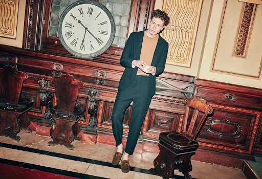 A stylish man leans against a wood-paneled wall with a large clock in a formal setting, wearing a dark suit and loafers.