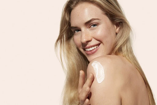 Close-up portrait of a smiling woman applying white cream to her bare shoulder against a neutral background.