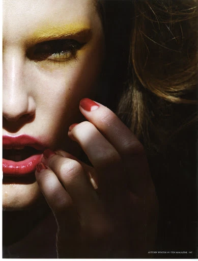 Close-up beauty shot features a woman with yellow eyeshadow and bright red lipstick, with her hand near her mouth in a dimly lit setting.