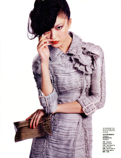 A woman with dark hair and red lipstick poses in a grey, ruffled dress with a clutch in a studio shot on a white background.