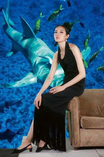 Glamorous woman in black dress sits on a couch in front of an underwater scene featuring sharks and tropical fish in ocean.