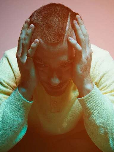 Close-up portrait of an adult man with hands on his head wearing a yellow sweater against a studio background with orange and blue gel lighting.