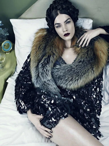 High-angle shot of a woman with dark hair and red lipstick reclining in bed wearing a fur stole and a dark feathered jacket.