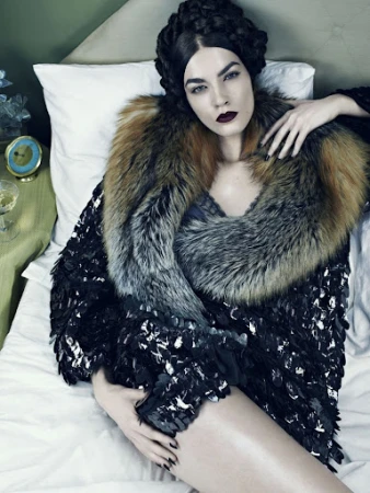 High-angle shot of a woman with dark hair and red lipstick reclining in bed wearing a fur stole and a dark feathered jacket.