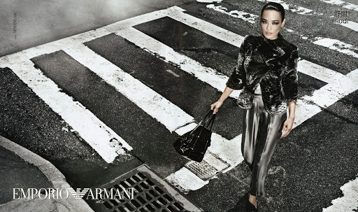 A woman stands on a crosswalk in an Emporio Armani ad, wearing a black, faux-fur jacket and shiny silver pants, holding a black bag.