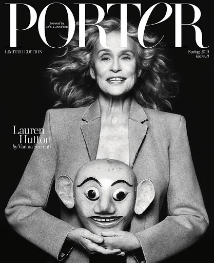 Monochrome portrait of Lauren Hutton holding a carved mask wearing a blazer against a dark background