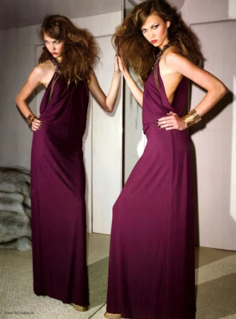 Model in long purple halter dress posing before a large mirror with voluminous brown hair and gold cuff bracelets