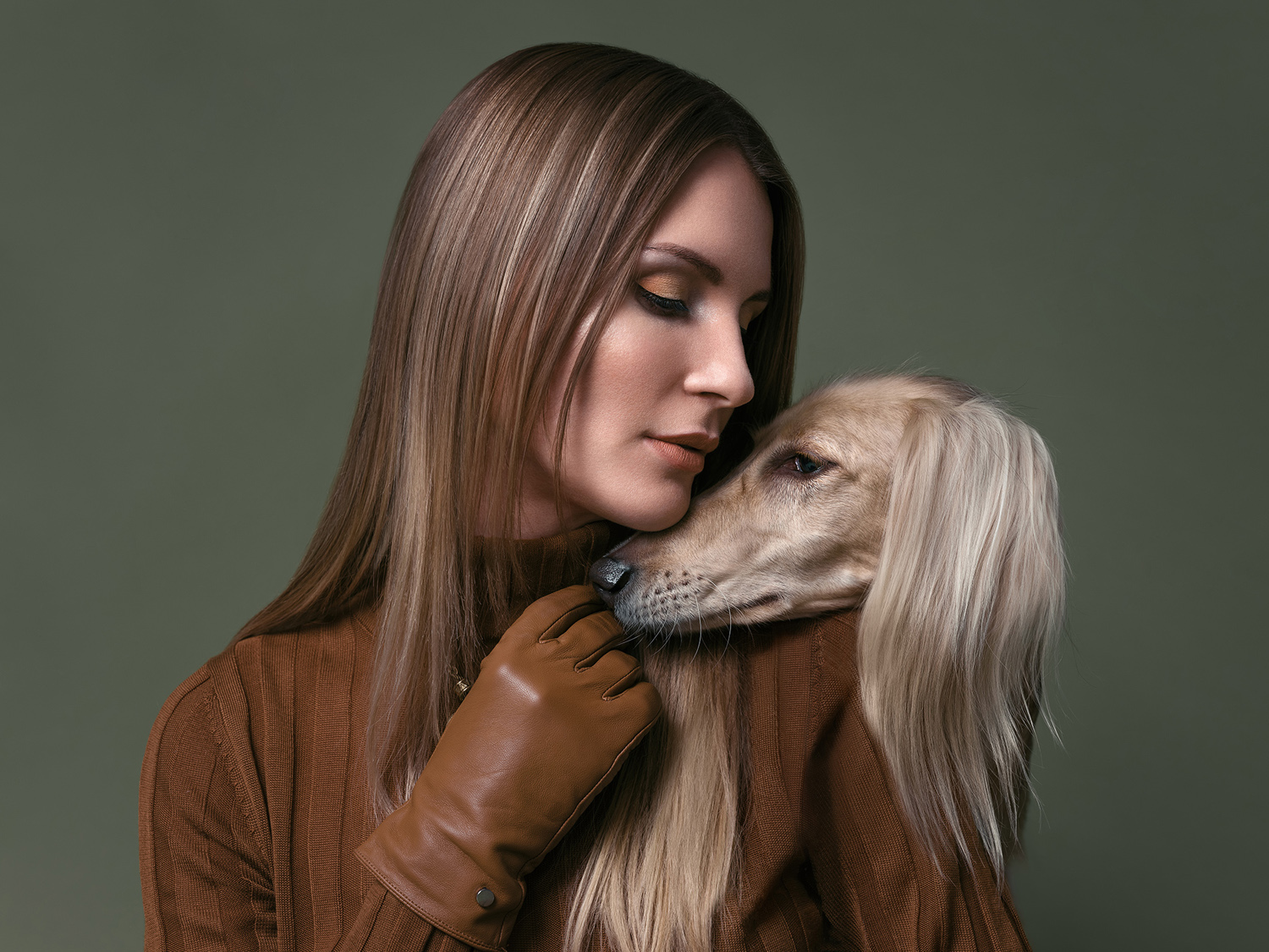 A blonde woman embraces a cream-colored Saluki dog, both indoors against a muted green backdrop in a medium studio shot.