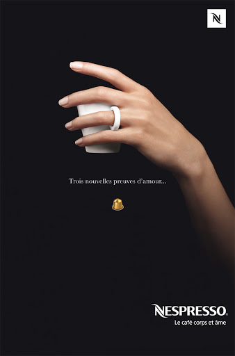 Coffee advertisement featuring a hand holding a white cup with a ring on the finger and a coffee pod on a dark background.
