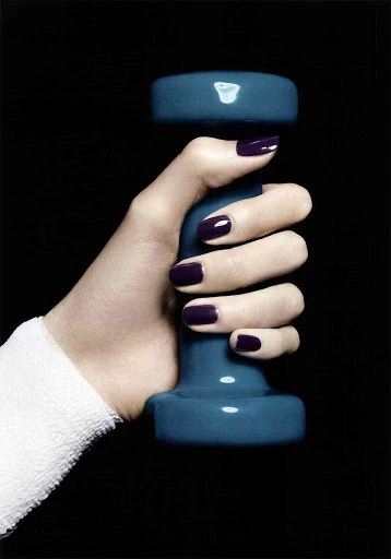 Hand holds blue dumbbell with dark purple manicure against black. Arm has a white wristband. Fitness and beauty shot.