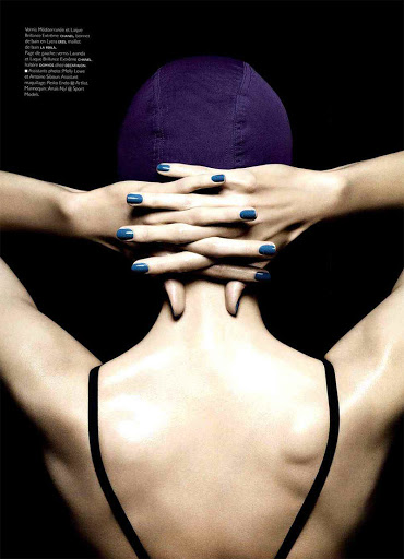 Back view of a woman with a swimming cap, hands interlaced behind her head and wearing a black top against a dark background.
