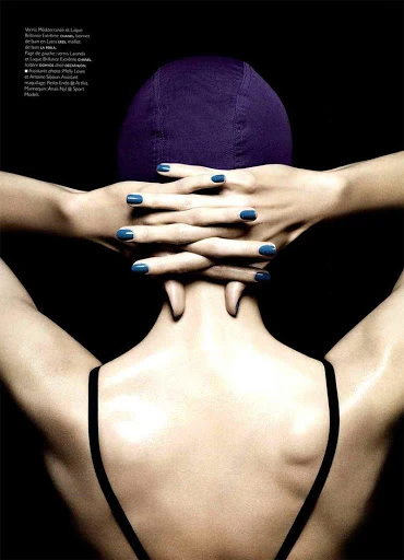 Back view of a woman with a swimming cap, hands interlaced behind her head and wearing a black top against a dark background.