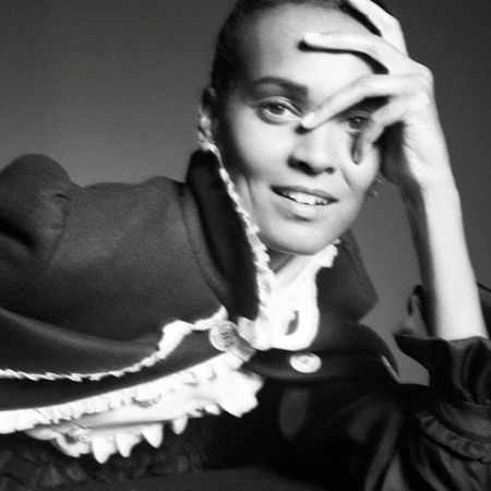 Portrait of a woman with a hand on her forehead, wearing a formal jacket with a ruffled collar against a neutral grey backdrop.