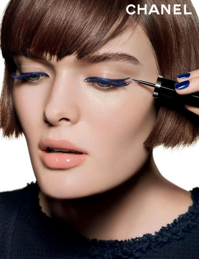 Beauty shot of a brunette woman with blue eyeliner. Makeup artist applying blue liner with a thin brush to her upper eyelid.