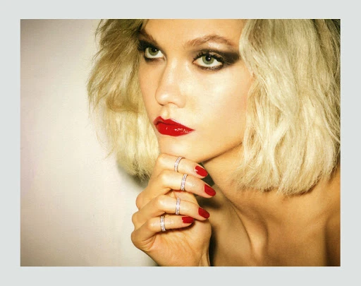 Close-up portrait of a blonde model with red lipstick, dark eye makeup, and bright red manicured nails wearing diamond rings.