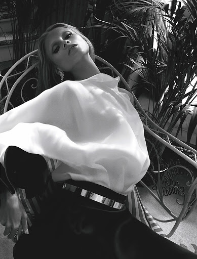 Black-and-white fashion portrait of a slender adult woman in a white blouse, sitting in a chair surrounded by tropical plants.