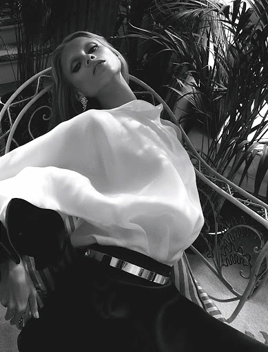 Black-and-white fashion portrait of a slender adult woman in a white blouse, sitting in a chair surrounded by tropical plants.