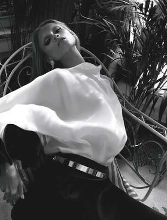 Black-and-white fashion portrait of a slender adult woman in a white blouse, sitting in a chair surrounded by tropical plants.