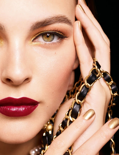 Close-up of a woman's face showcasing dark red lipstick, gold eyeshadow, and matching gold nail polish against a black background.