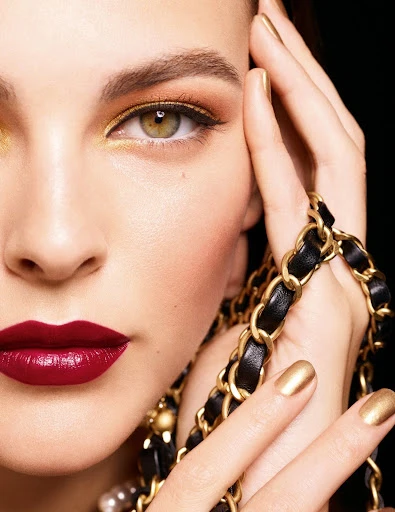 Close-up of a woman's face showcasing dark red lipstick, gold eyeshadow, and matching gold nail polish against a black background.