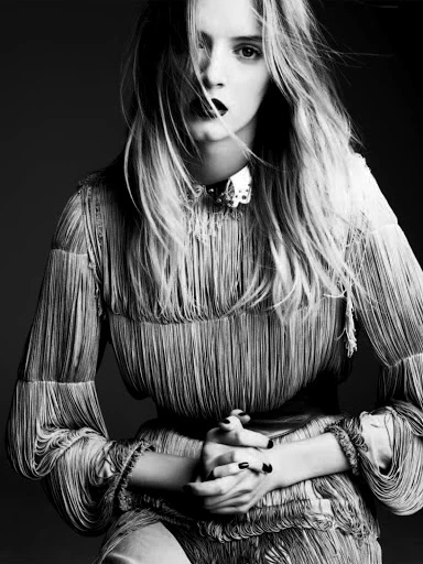 A portrait of a fair-skinned woman in a fringe dress with long blonde hair and dark lipstick, with her hands folded in front of her.