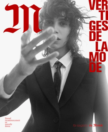 Black and white editorial portrait of a person with dark curly hair wearing a suit and tie, reaching toward the camera.