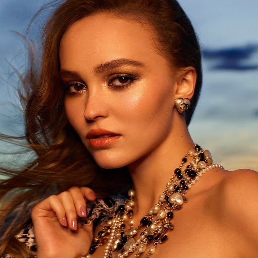 Close-up portrait of a woman with wavy brown hair, wearing Chanel heart earrings and layered pearl necklaces against a diffused background.