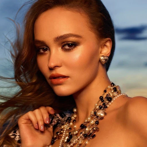 Close-up portrait of a woman with wavy brown hair, wearing Chanel heart earrings and layered pearl necklaces against a diffused background.