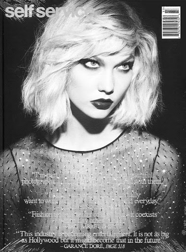 Monochrome magazine cover portrait of a blonde woman wearing a dark sheer crystal-embellished top.