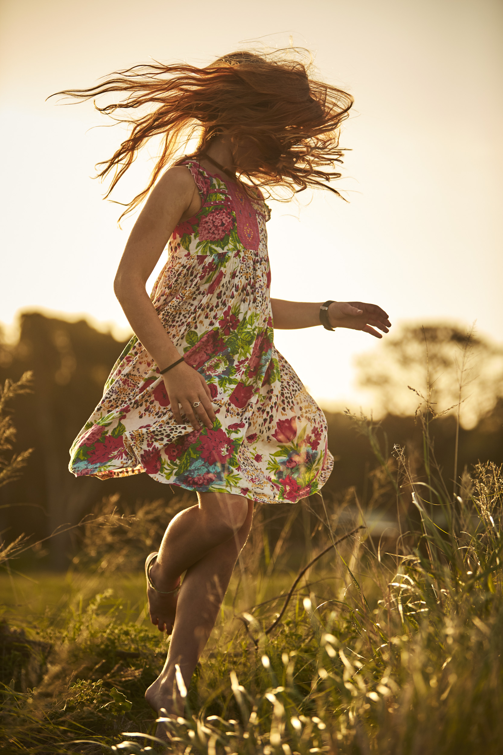 Young woman in a floral dress jumps barefoot in a grassy field at golden hour, hair flying, capturing a carefree, natural moment outdoors.