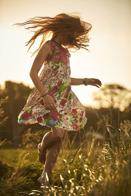Young woman in a floral dress jumps barefoot in a grassy field at golden hour, hair flying, capturing a carefree, natural moment outdoors.