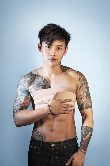 Portrait of a tattooed Asian man wearing jeans, posing shirtless with a beige bandage wrapped around his chest against a light blue background.