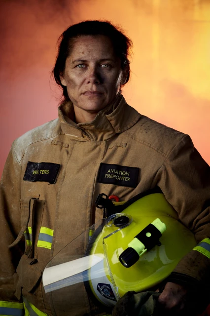 Portrait of a female aviation firefighter wearing a tan uniform and holding a helmet against a red and orange background.