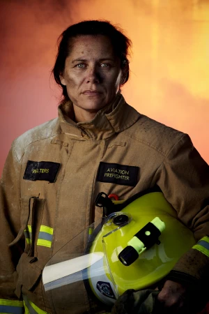 Portrait of a female aviation firefighter wearing a tan uniform and holding a helmet against a red and orange background.