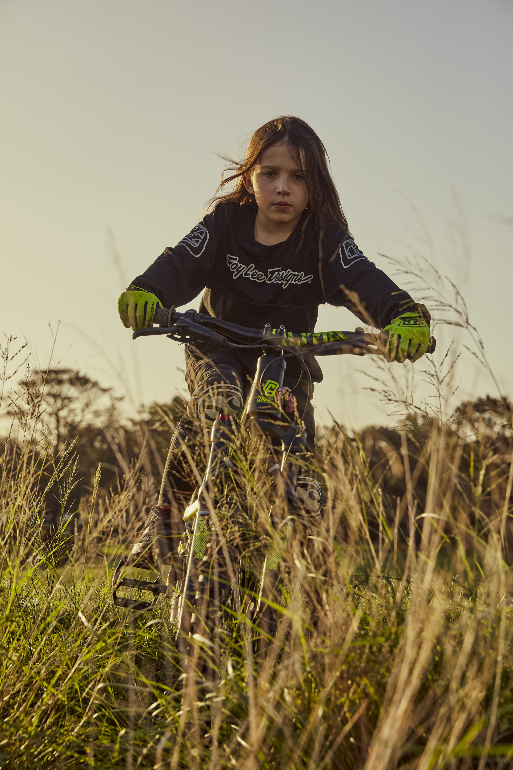 Young child riding a green and black mountain bike through a grassy field at sunset. The rider wears a black long sleeve shirt and green gloves.