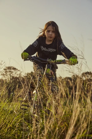 Young child riding a green and black mountain bike through a grassy field at sunset. The rider wears a black long sleeve shirt and green gloves.