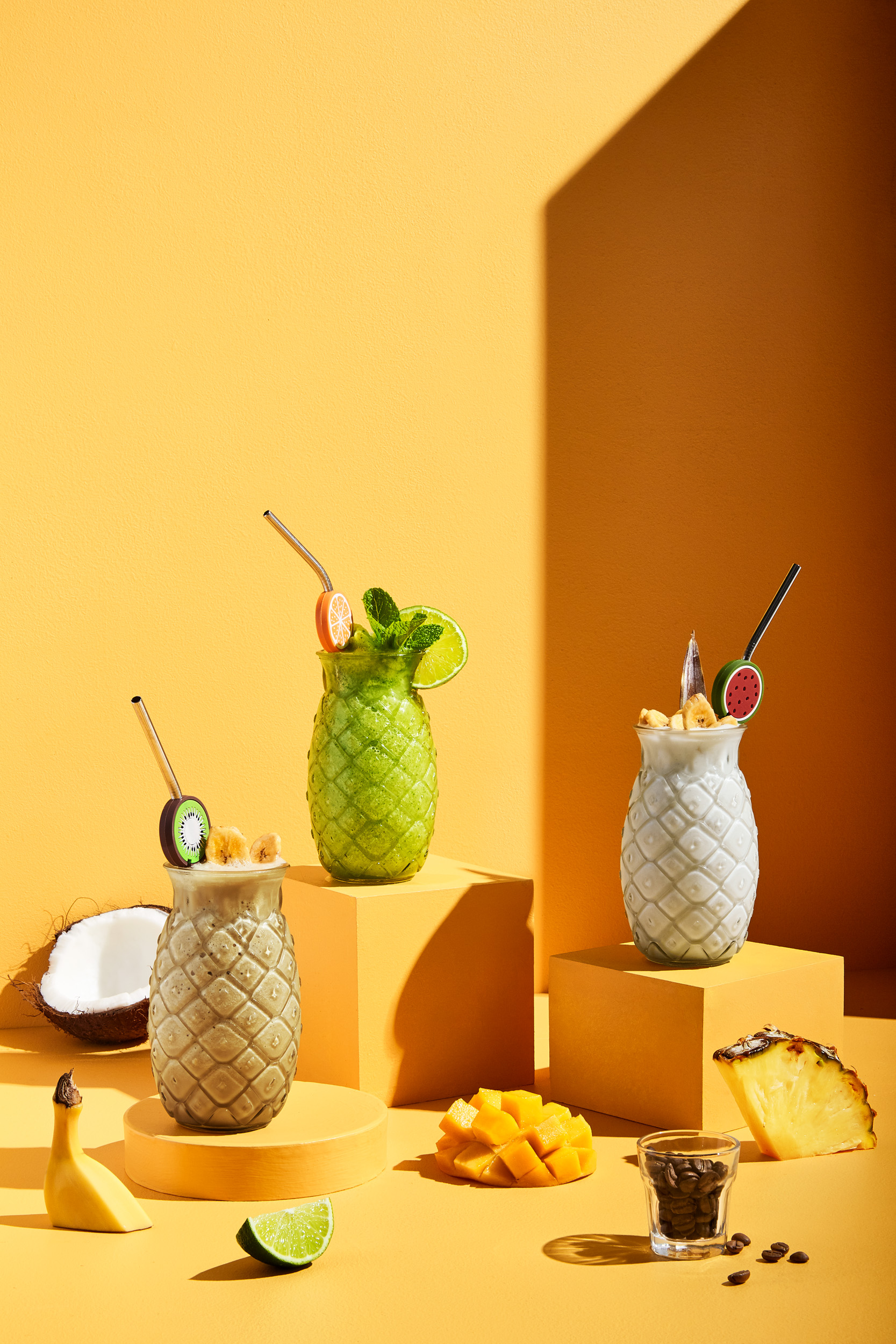 Three tropical fruit smoothies in pineapple-shaped glasses on orange platforms with mango, banana, lime, kiwi, and coffee beans.