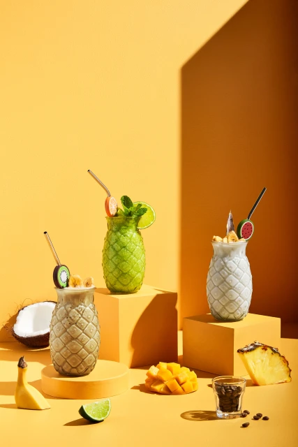 Three tropical fruit smoothies in pineapple-shaped glasses on orange platforms with mango, banana, lime, kiwi, and coffee beans.