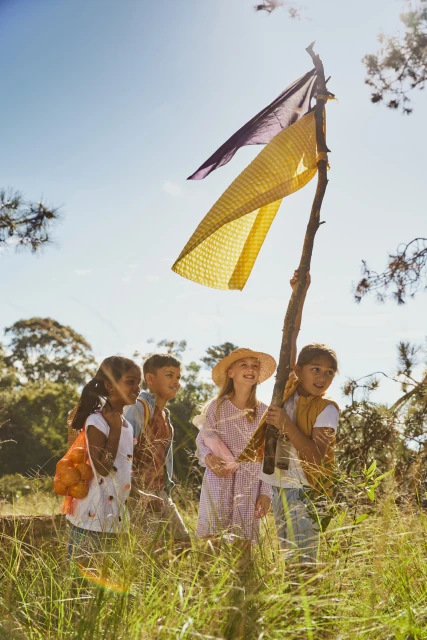 A group of five children outdoors hold up a stick with colorful fabric attached to it in a field of tall grass under a bright blue sky.