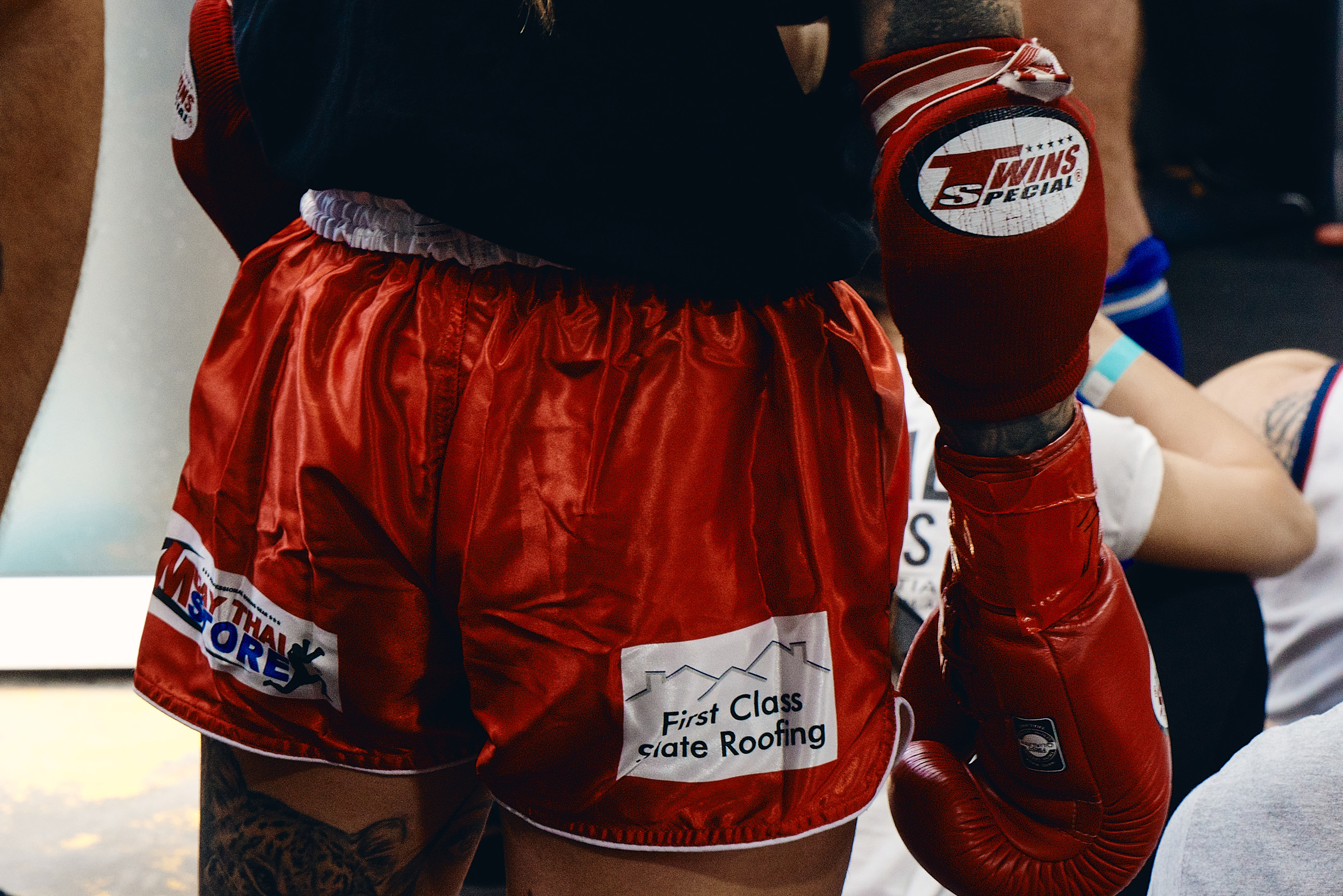 Close-up of a boxer in red boxing shorts and gloves, advertising local businesses. Visible tattoos and fight wear are prominent.