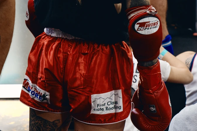 Close-up of a boxer in red boxing shorts and gloves, advertising local businesses. Visible tattoos and fight wear are prominent.