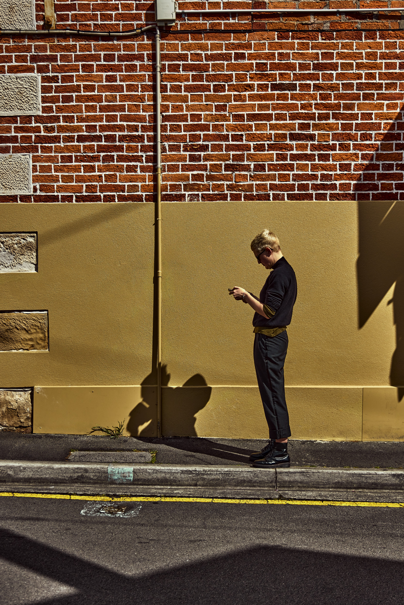 A young adult stands outside a brick building using a smartphone, captured in an urban setting during daylight with shadows.