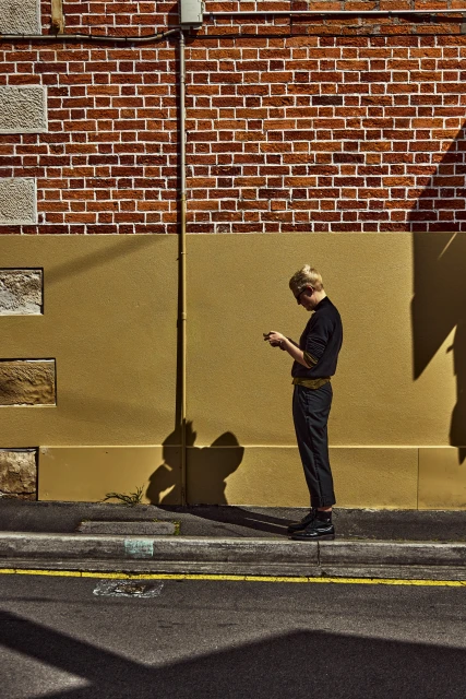 A young adult stands outside a brick building using a smartphone, captured in an urban setting during daylight with shadows.
