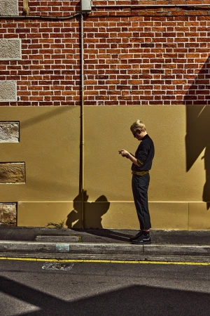 A young adult stands outside a brick building using a smartphone, captured in an urban setting during daylight with shadows.