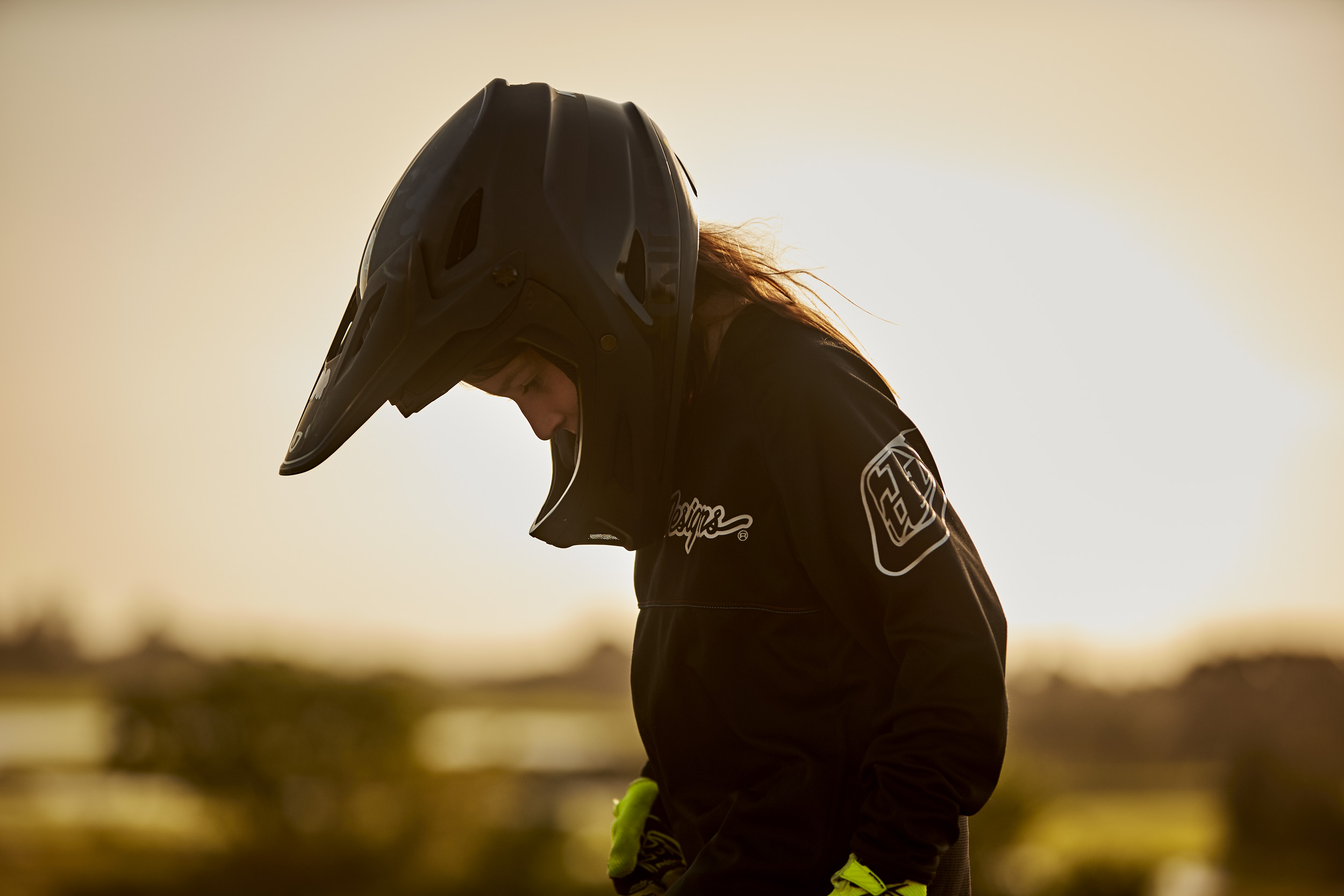 Teenage cyclist in black helmet and jersey looks down in a rural setting during golden hour, showcasing athletic apparel.